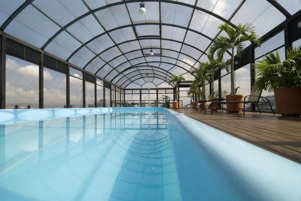 Indoor swimming pool with arched translucent roof, palm trees, and outdoor seating.
