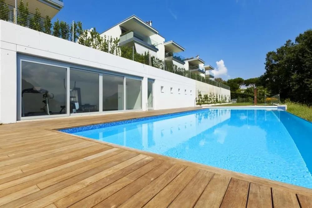 Modern white building with a pool and wooden deck, overlooking blue water.