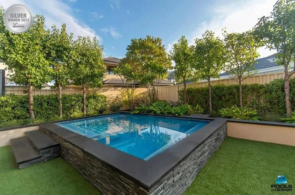 Small backyard with a square pool surrounded by stone and artificial turf, trees in the background.