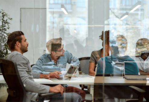 People in a meeting room, visible through glass. They are seated around a white table, some with laptops.