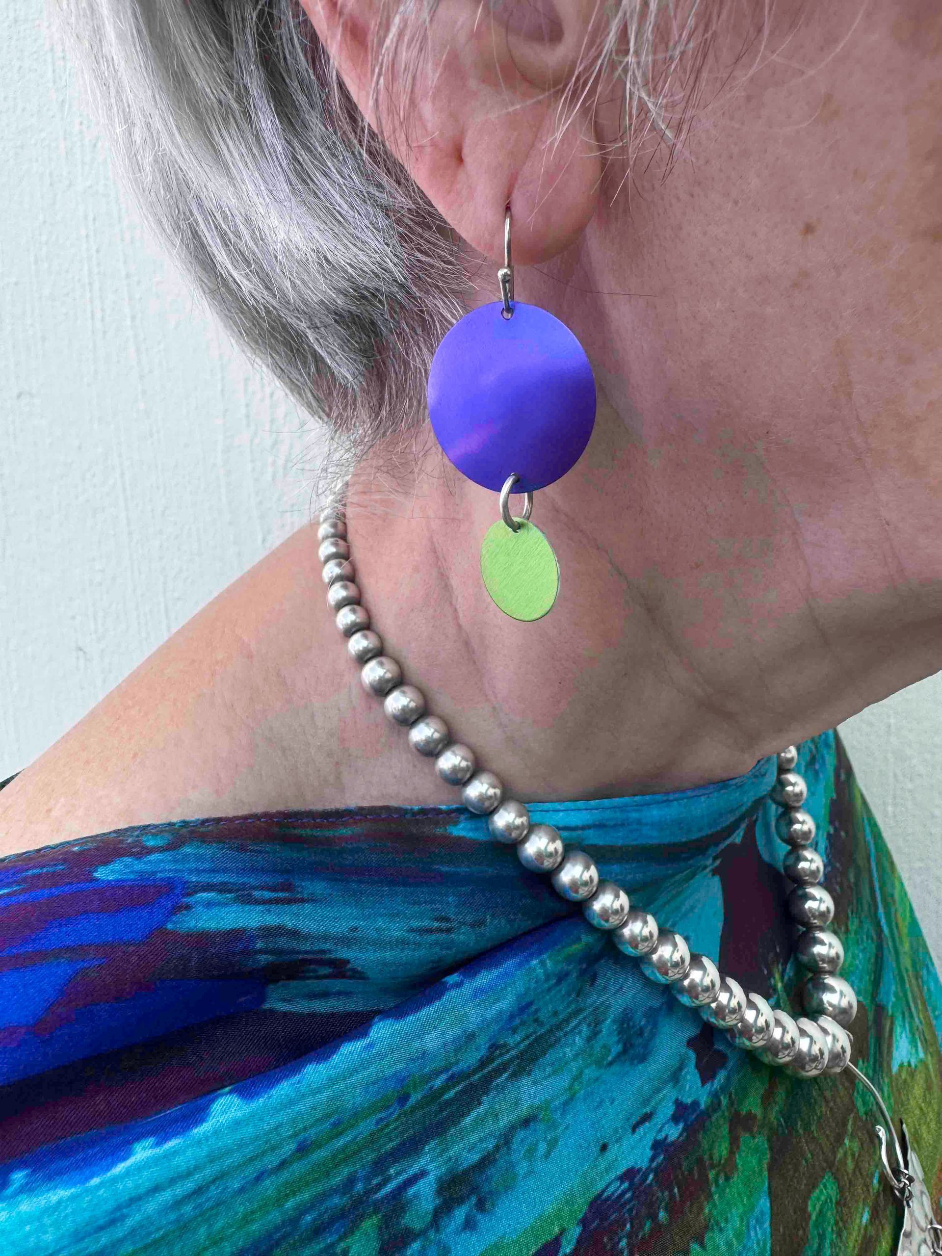 Woman wearing purple and green earrings, silver beaded necklace, and patterned blue scarf.
