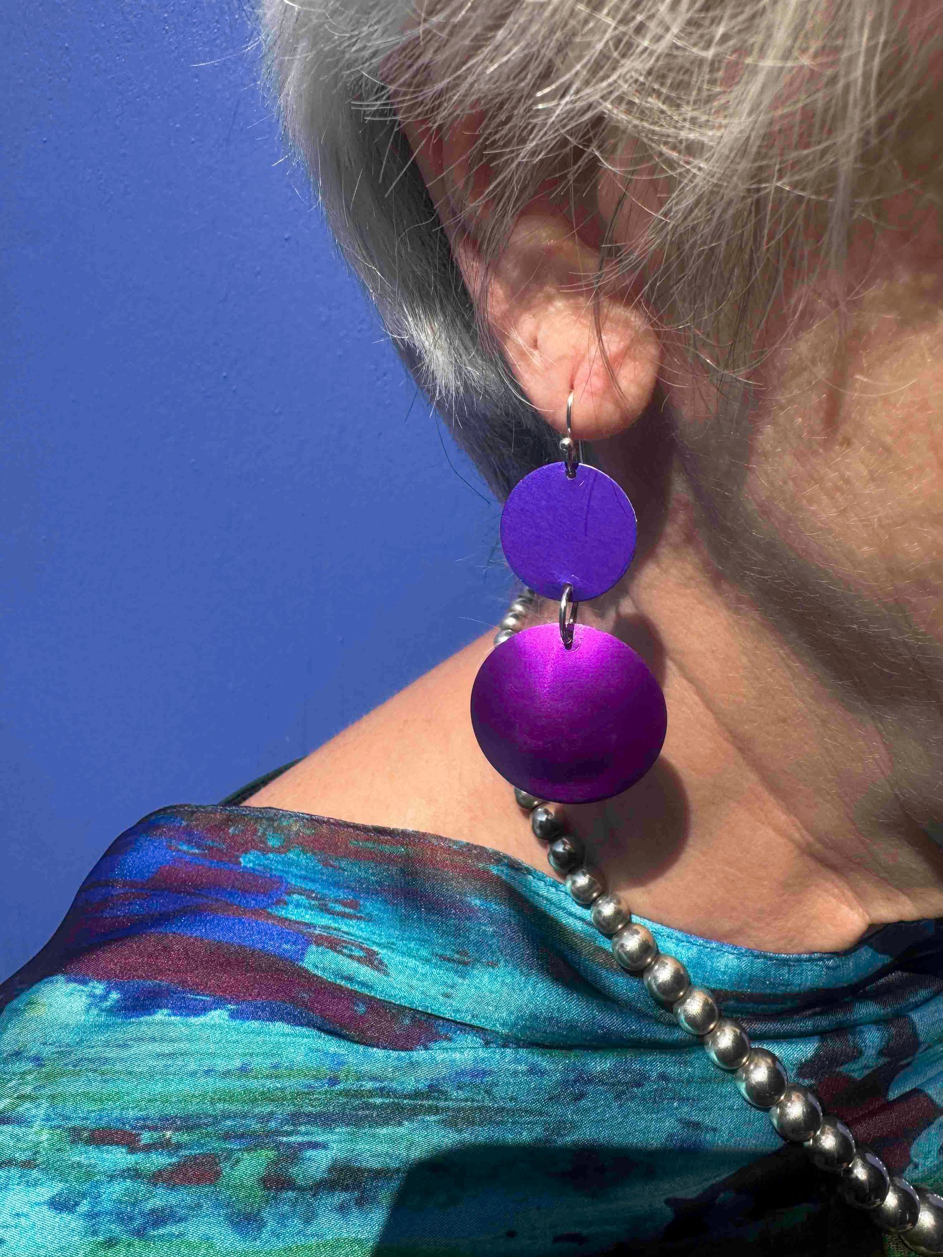 Person wearing purple and silver earrings, with blue and teal patterned scarf, against a blue background.