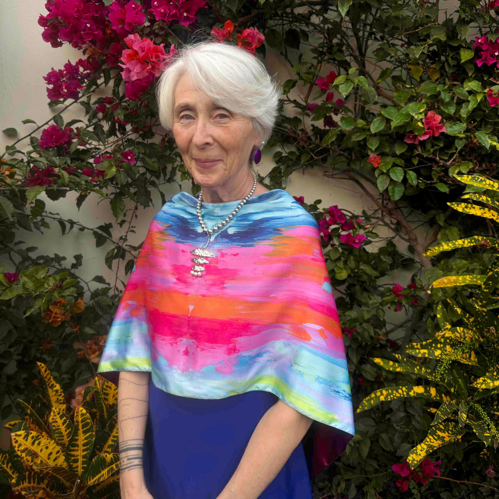 Woman with white hair wearing a colorful shawl stands in front of bougainvillea.