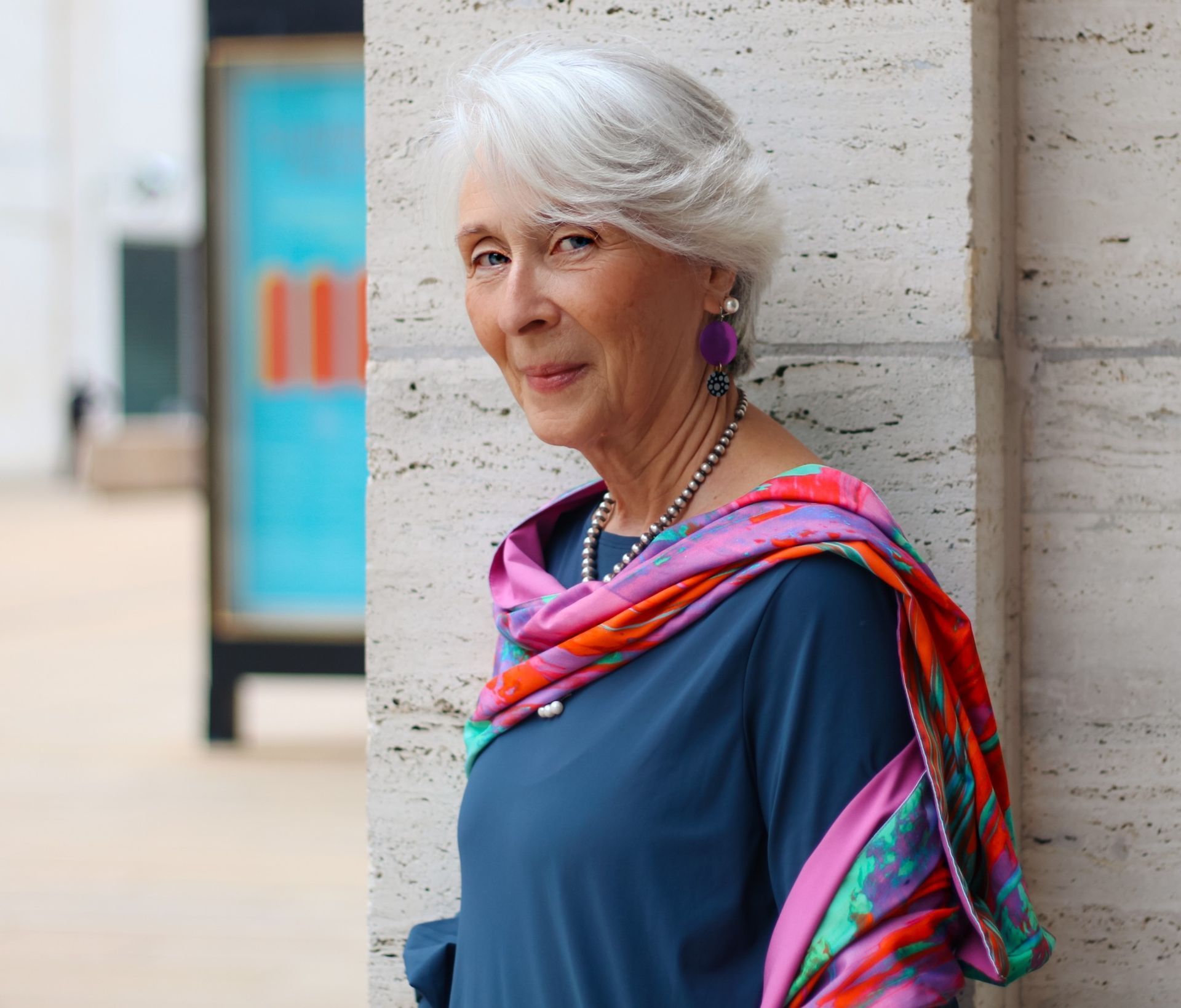 Woman with gray hair, wearing a scarf and earrings, leaning against a building.