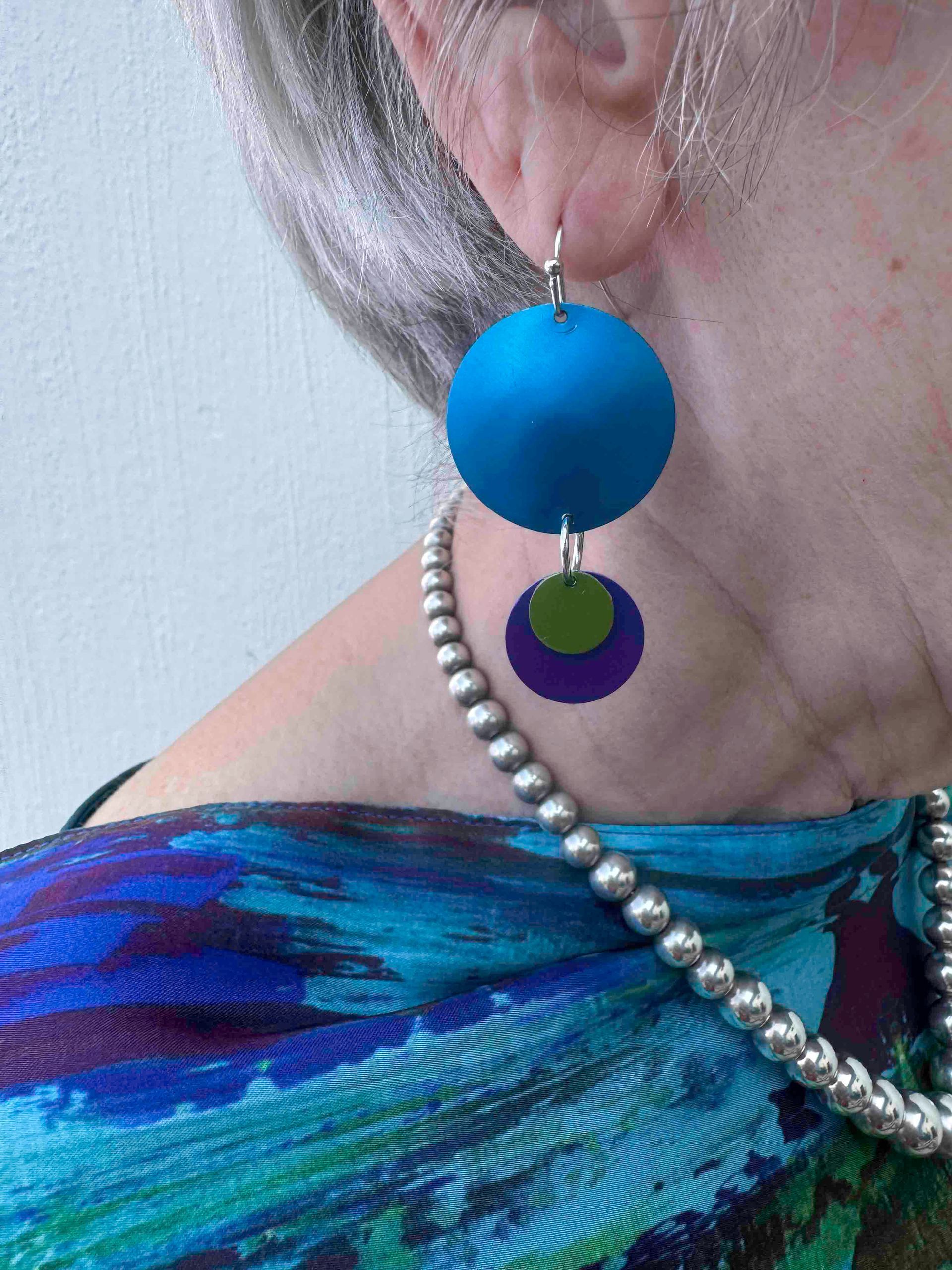 Close-up of a person's ear wearing blue and purple spherical earrings. A beaded necklace and a patterned teal top are also visible.