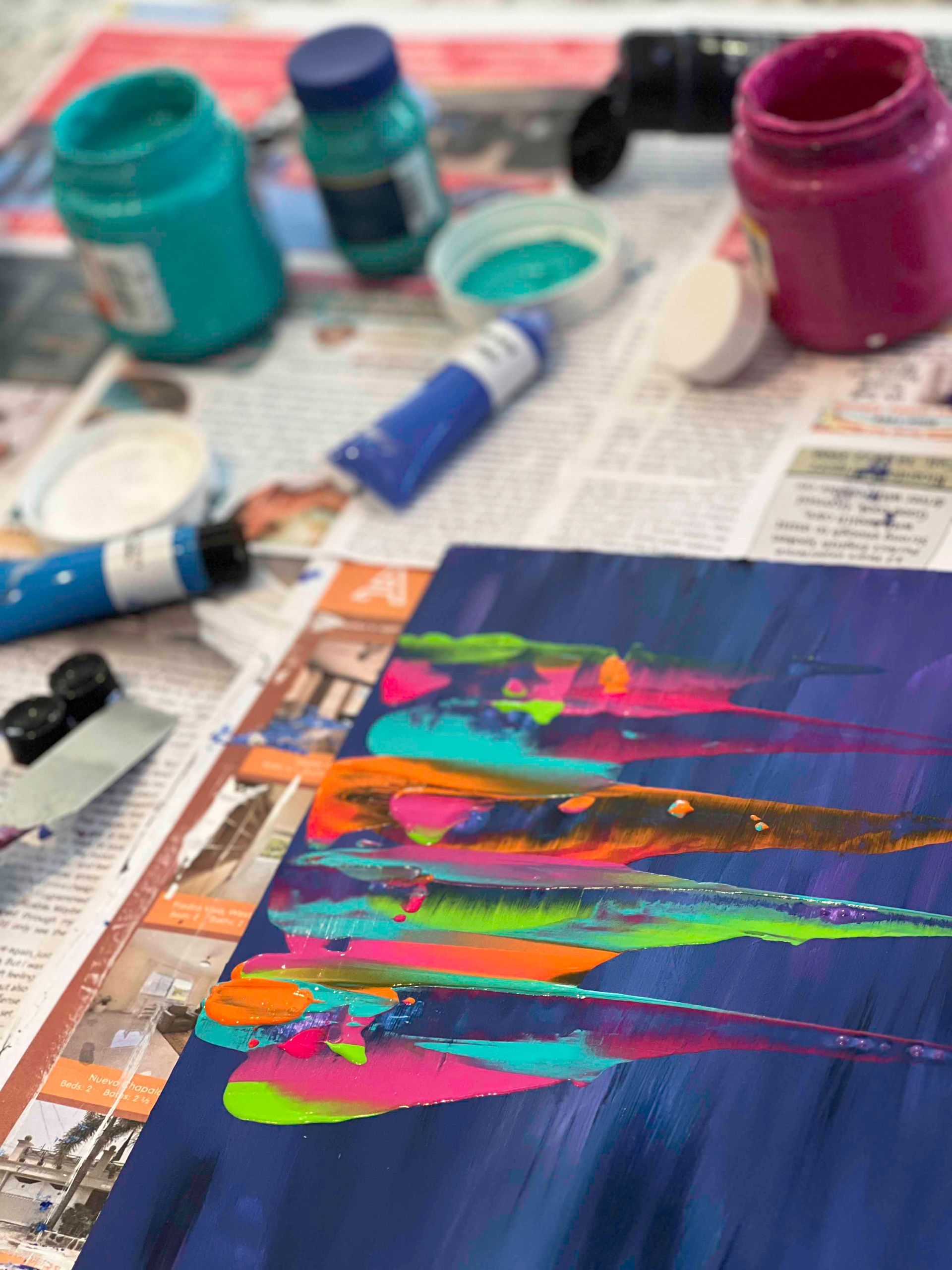 Painting in progress with vibrant streaks of color against a dark blue background; paint supplies nearby.