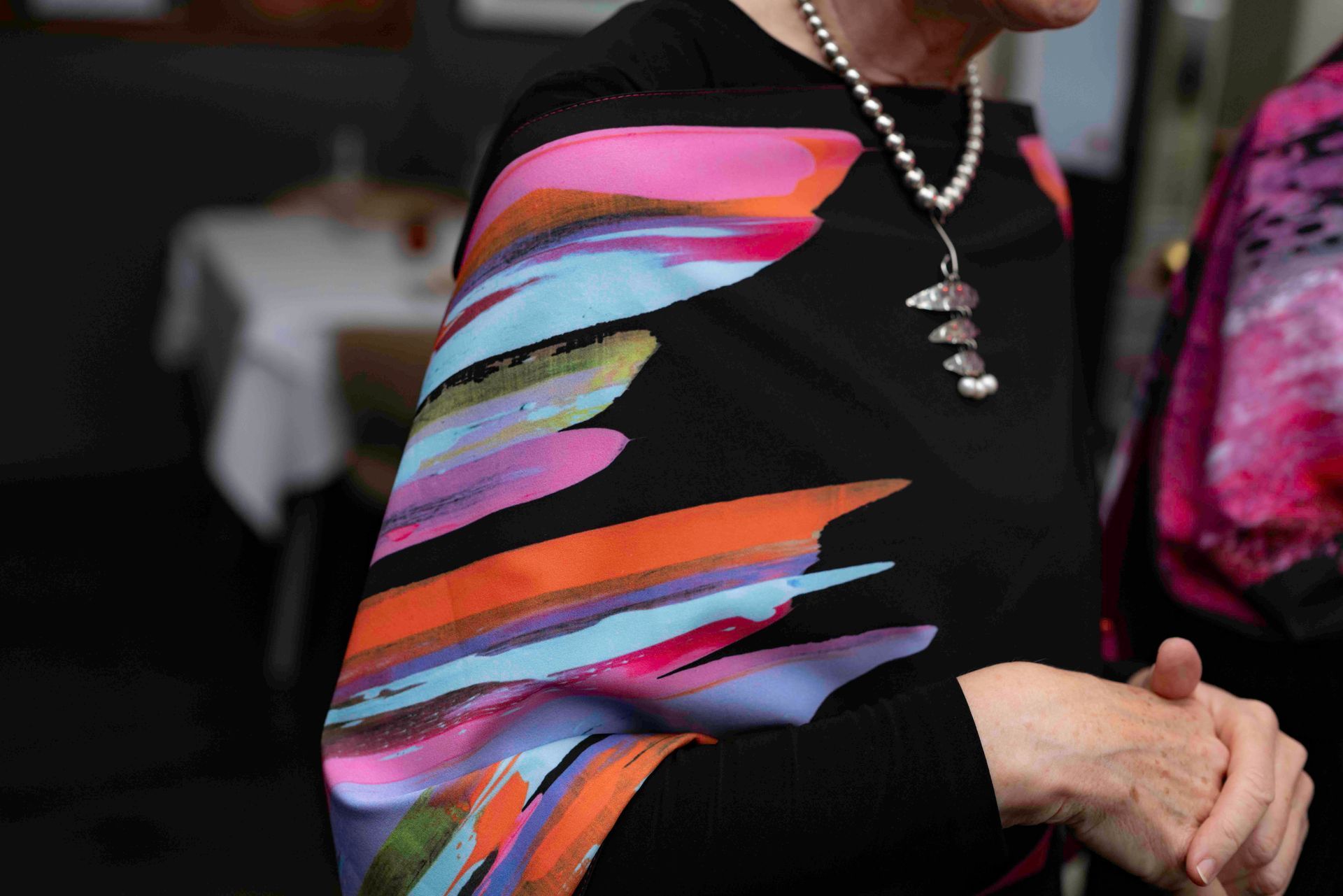 Woman wearing a black top with colorful abstract sleeve design, pearl necklace, arms crossed.