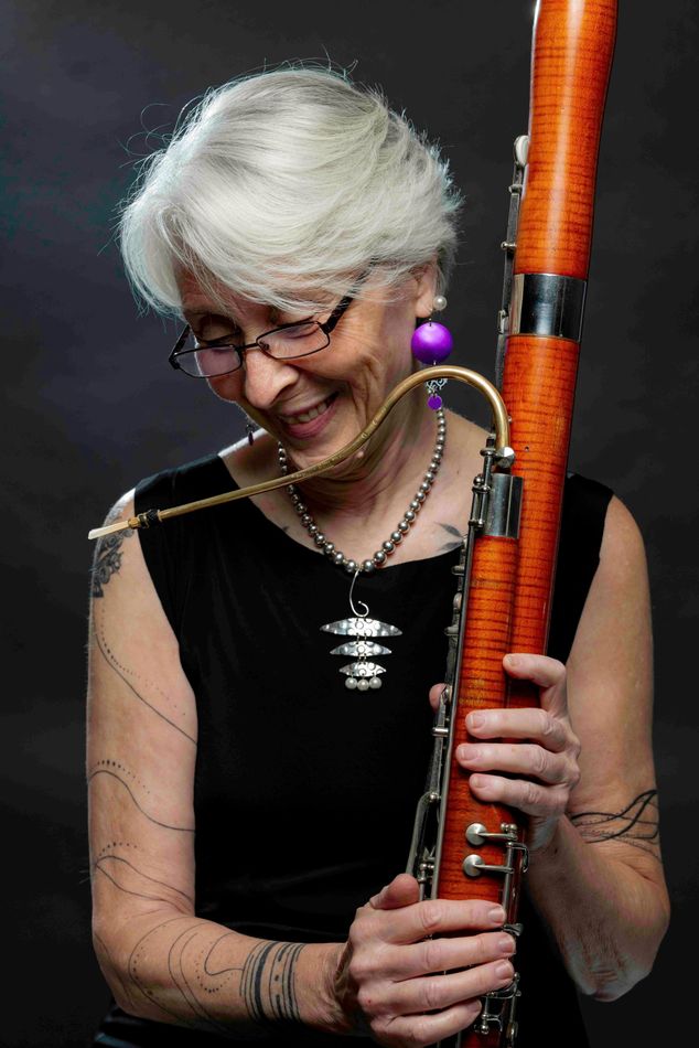 Woman holding a bassoon, smiling. Black sleeveless top, silver jewelry, glasses, gray hair. Dark background.