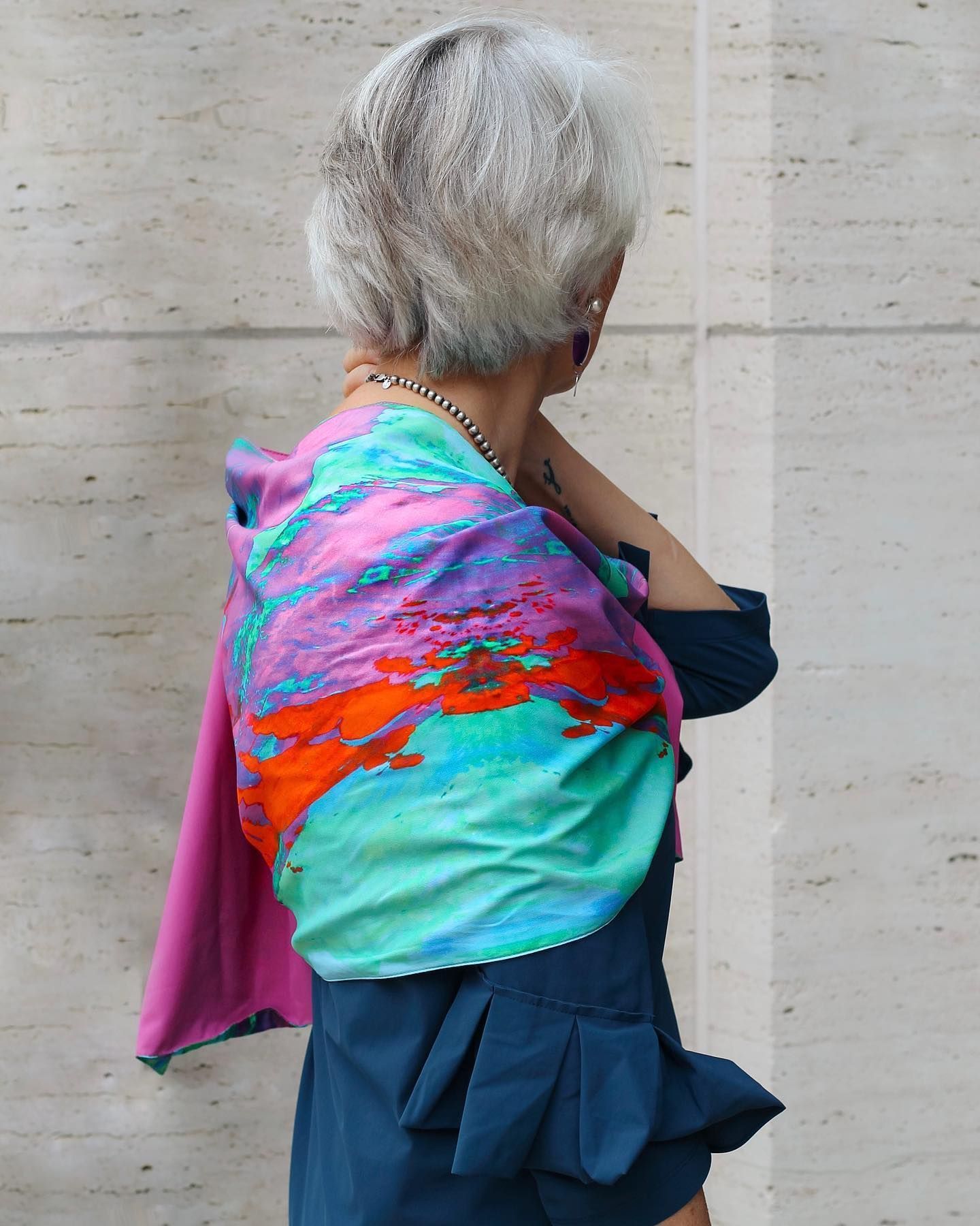 Person in blue dress, wearing a colorful shawl. Silver hair, standing in front of a beige wall.