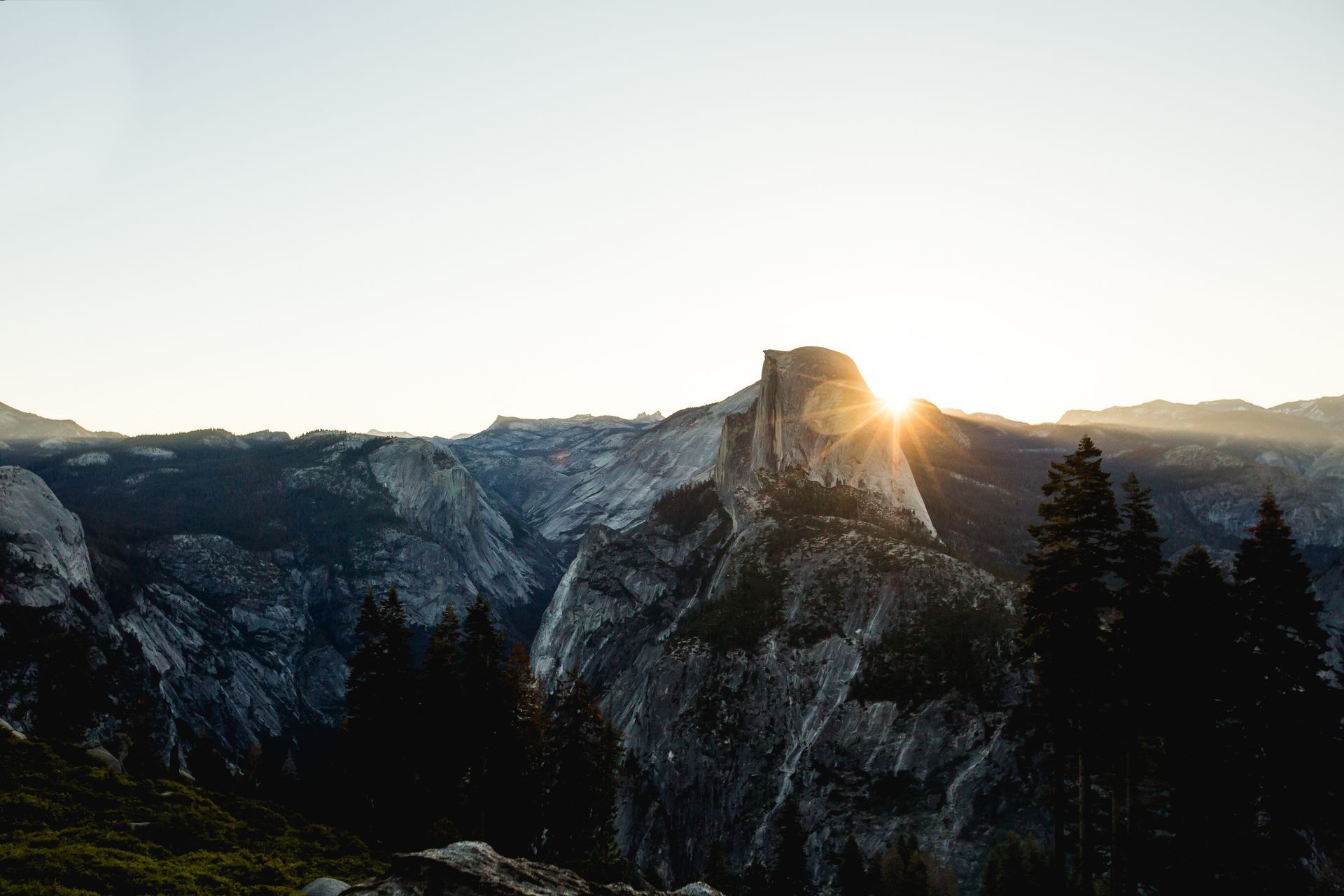 Sun sets behind Half Dome in Yosemite Valley, casting a golden glow on the mountain and trees.