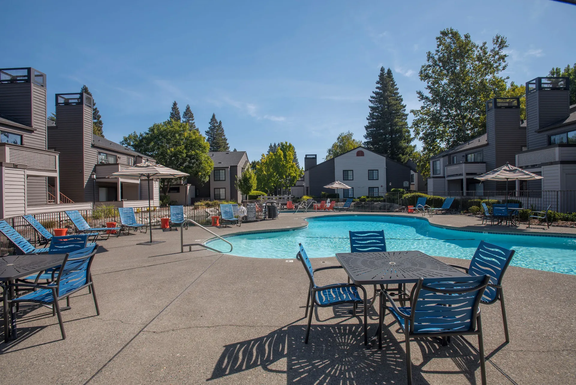 Pool area with tables, chairs, and lounge chairs. Apartment buildings and trees in the background. Blue sky.