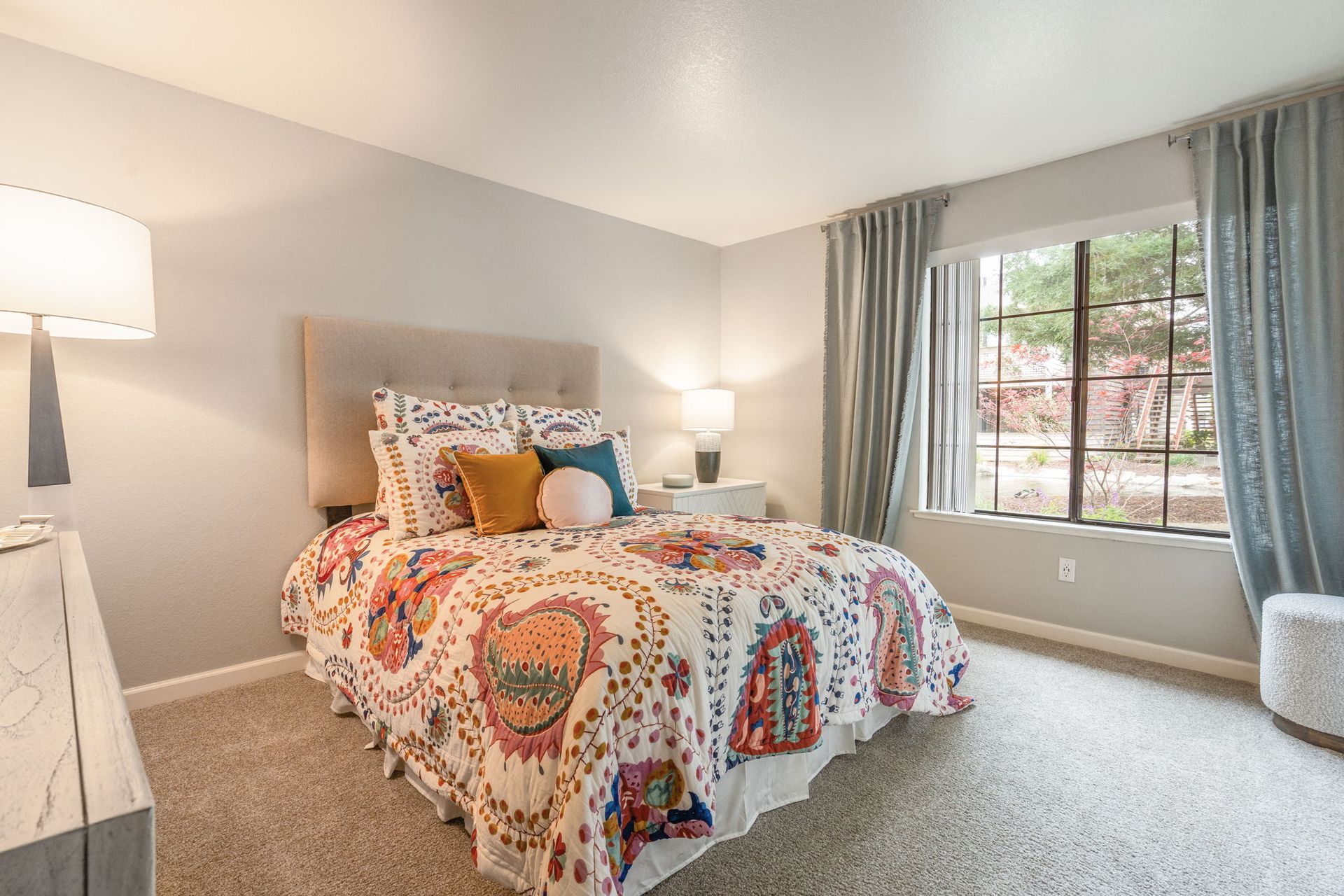 Bedroom in apartment with queen bed, colorful quilt, two nightstands, and large window with blue curtains.