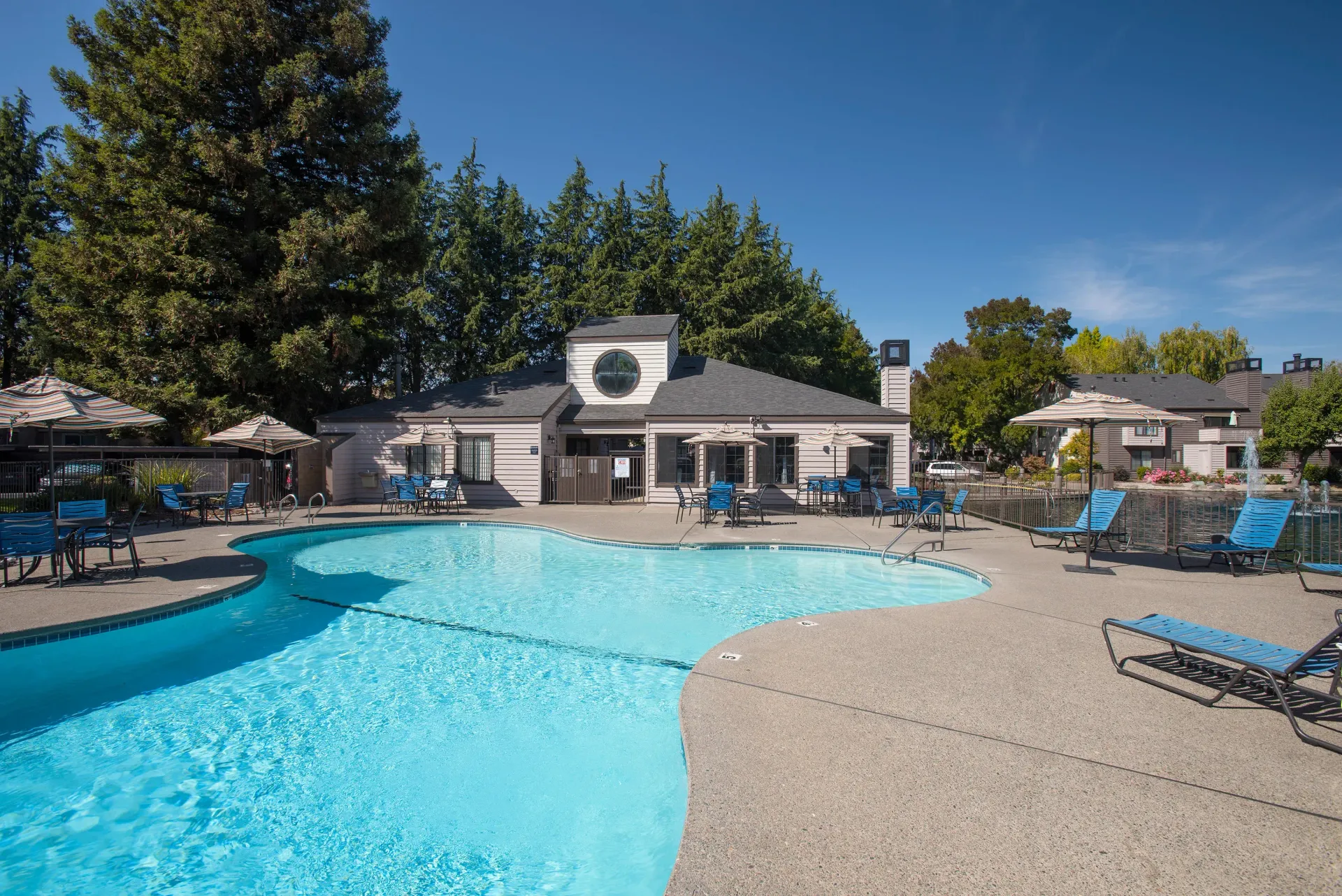 Swimming pool with lounge chairs, building, and trees on a sunny day.