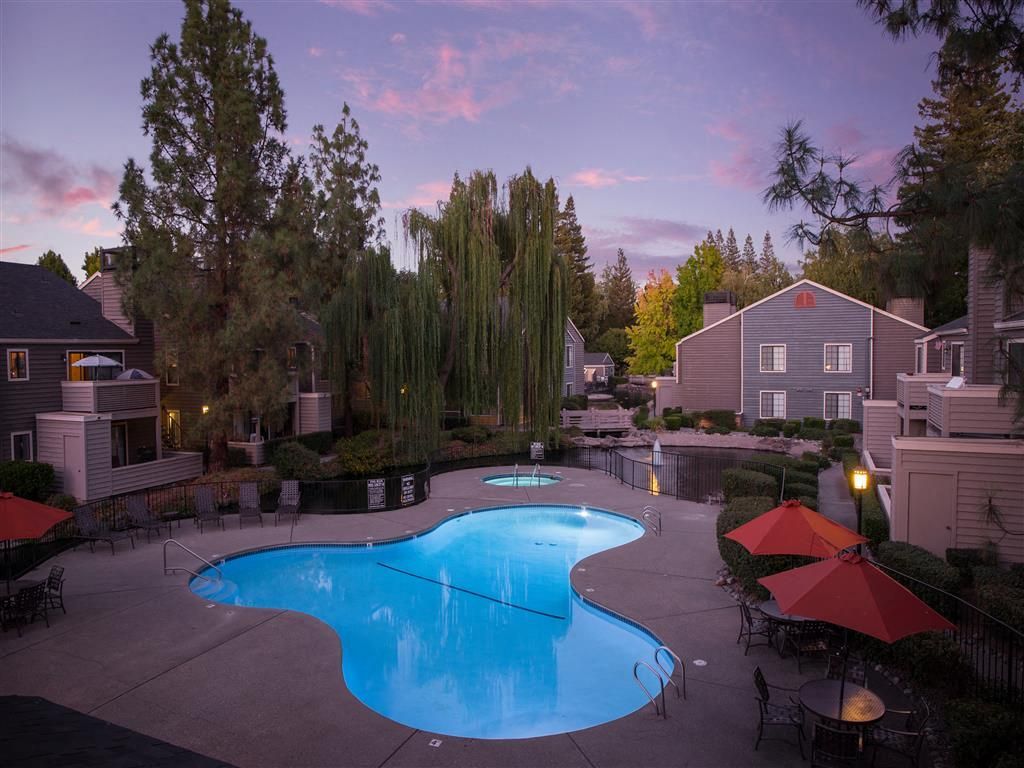 Outdoor pool area of an apartment community at dusk with lounge chairs and red umbrellas.