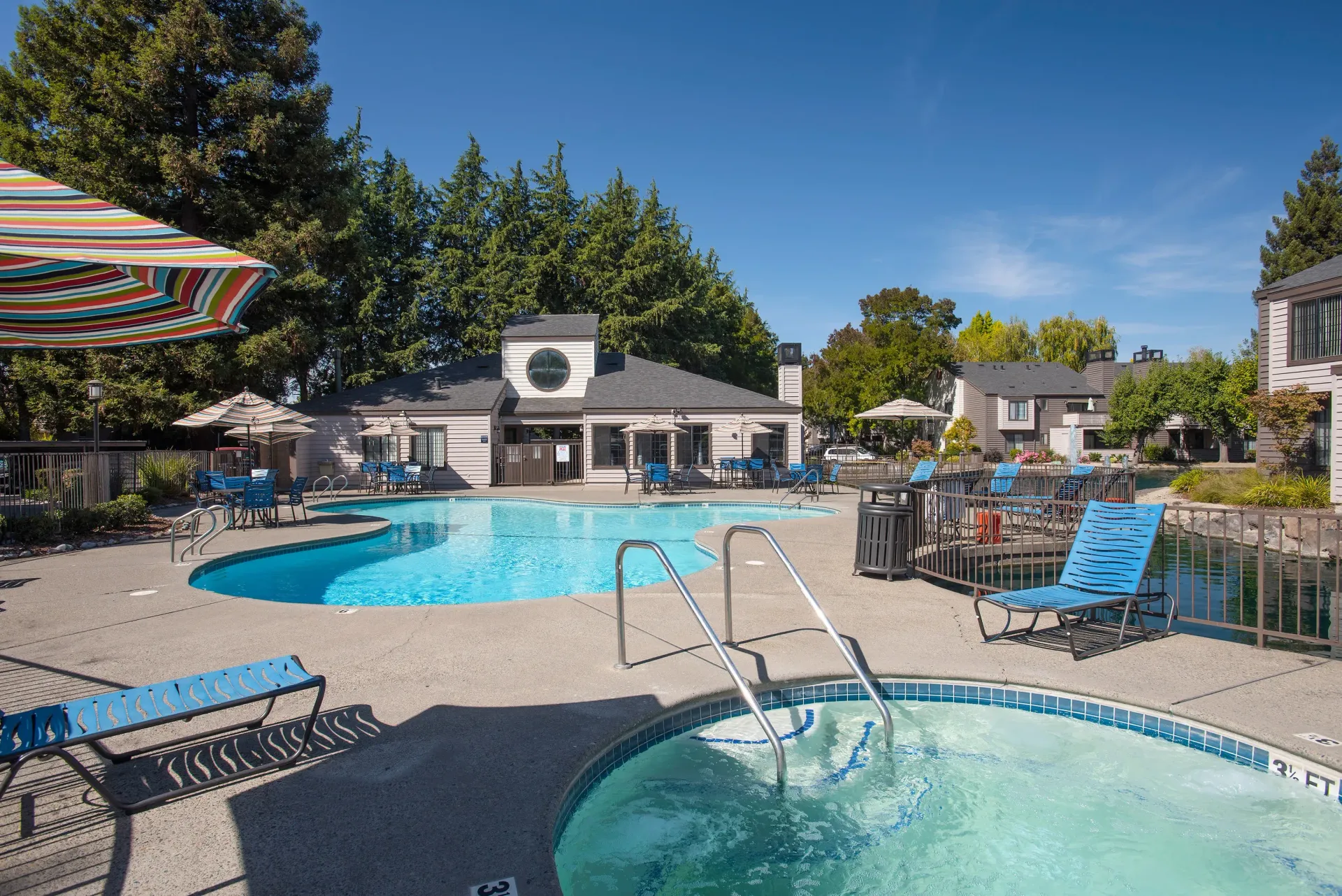 Pool and jacuzzi at an apartment complex with blue lounge chairs and umbrellas.