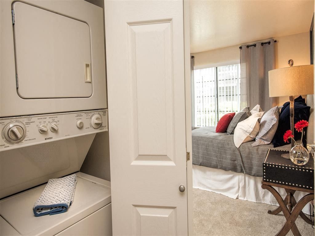 Stacked washer and dryer in a closet beside a bedroom with a bed and nightstand