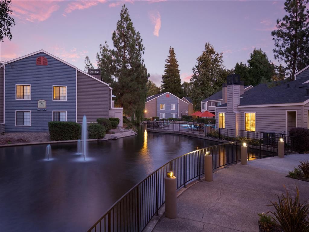 Exterior view of a community courtyard with a pond, fountains, a railing walkway, and lit buildings at dusk.