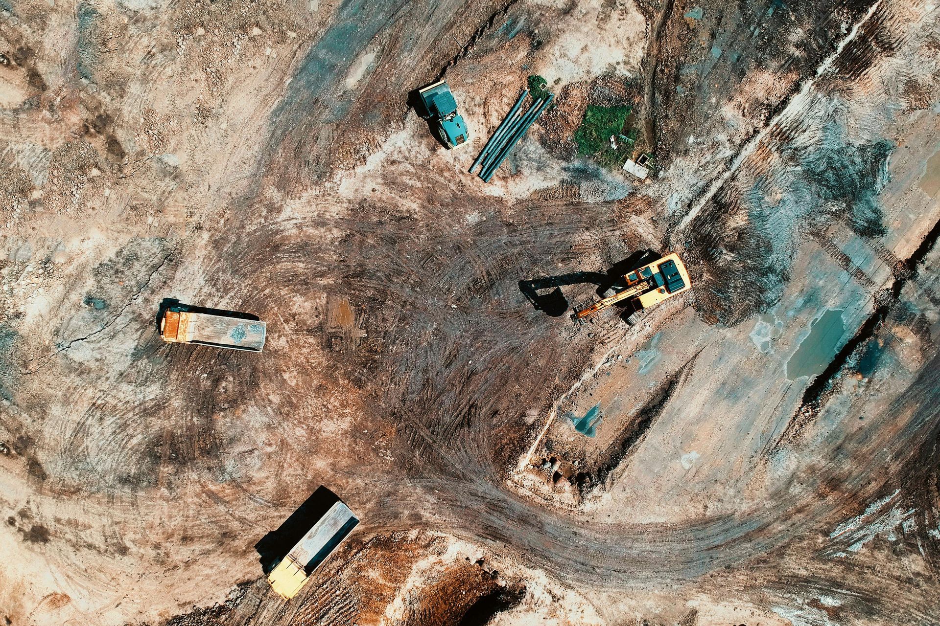 An aerial view of a construction site featuring an excavator and two dump trucks on dirt ground.