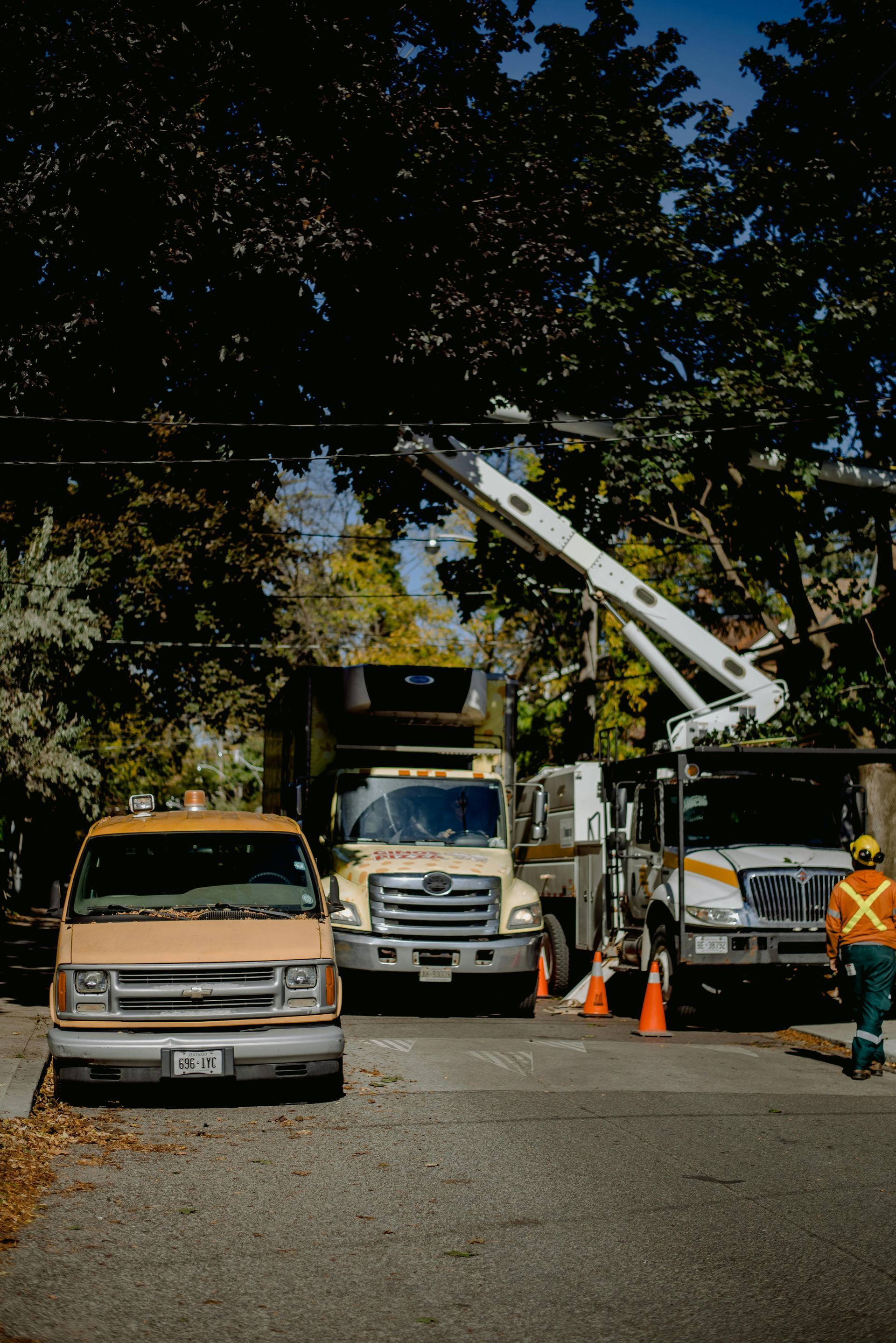 A utility bucket truck, a dump truck, and a van parked on a residential street with a worker in safety gear nearby.