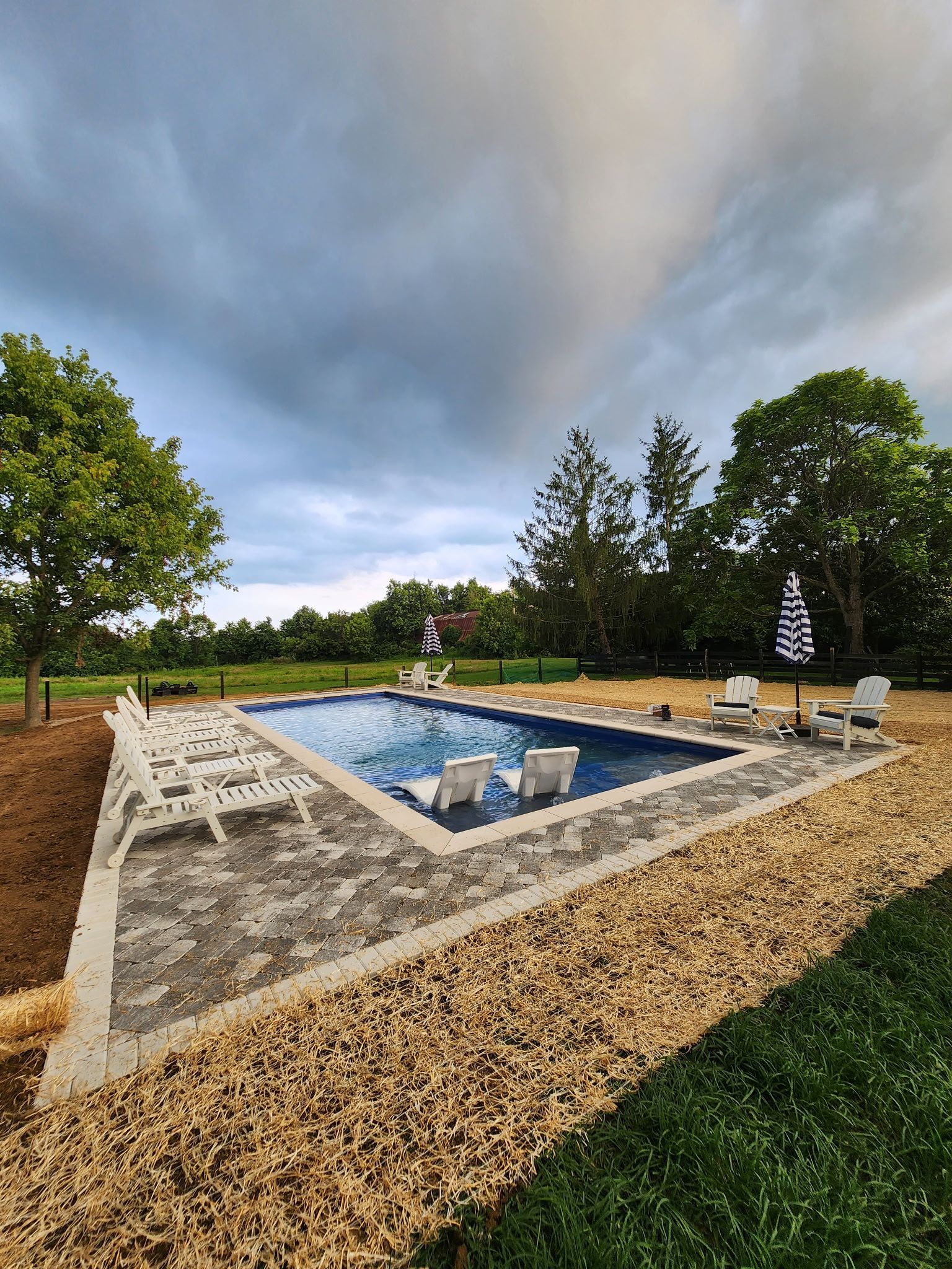 A rectangular pool with light blue water, surrounded by gravel and a concrete border, under a cloudy sky.
