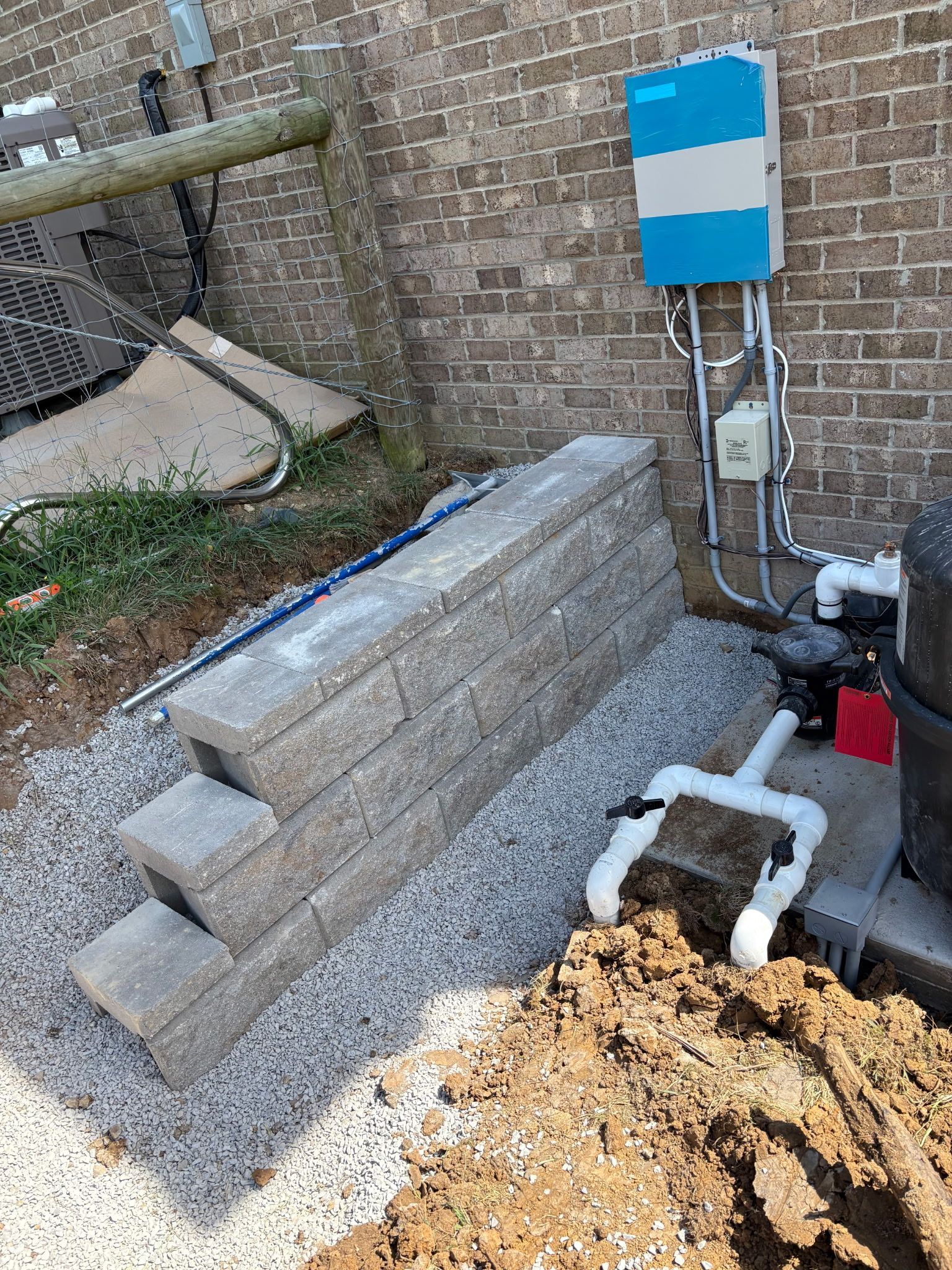 Retaining wall made of gray blocks near pool equipment against a brick wall.
