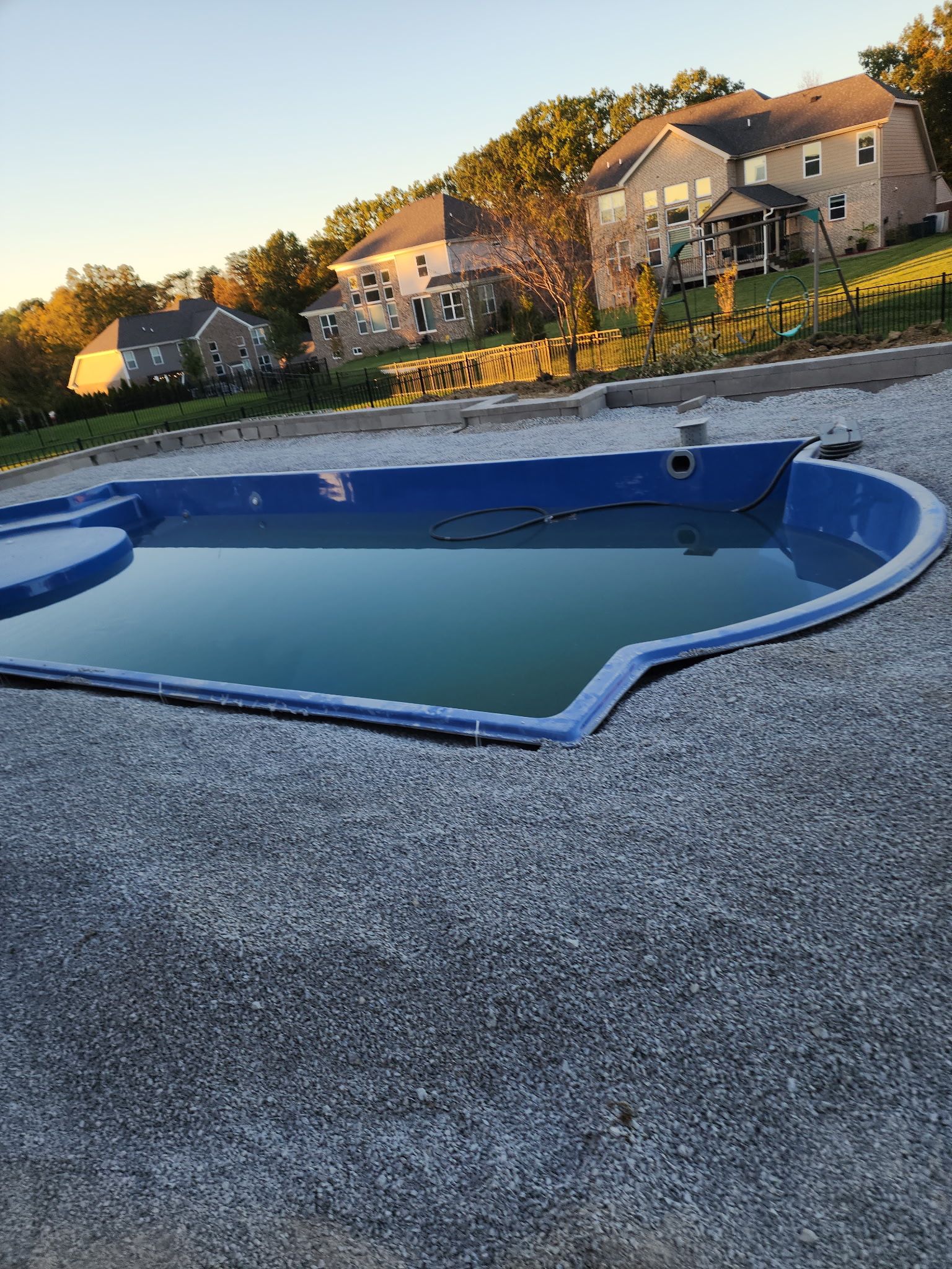 Empty blue swimming pool surrounded by gravel in a residential area. Houses and trees are in the background.