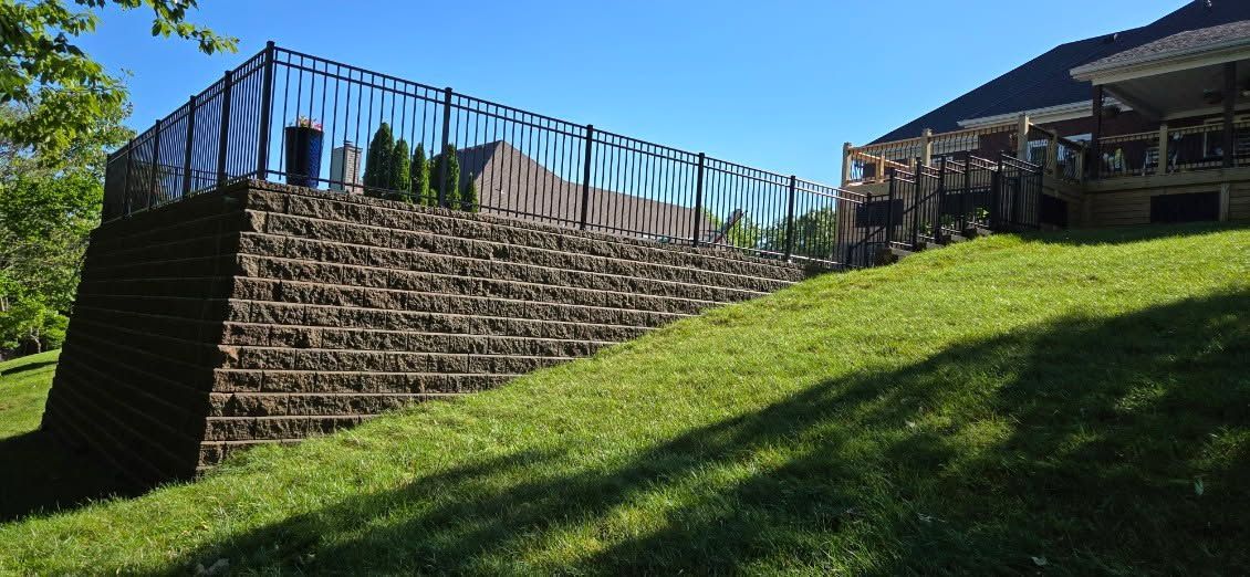 Stone retaining wall with black railing beside a grassy hillside under a blue sky