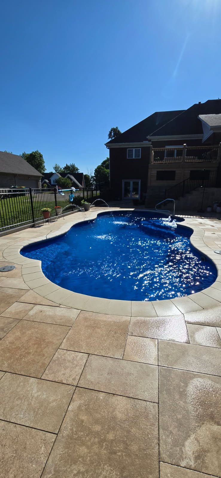 A freeform swimming pool surrounded by beige tiles, with a house and clear blue sky in the background.