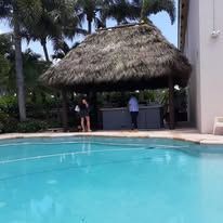 Tiki hut by a pool. Two people stand near a counter. Palm trees in the background. Blue pool water.