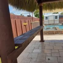 Wooden bar under a thatched roof with a view of a pool, fence, and brick patio.