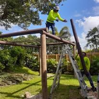 Two workers building a wooden structure outdoors; one on a ladder, another on the ground.