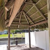 Palm-thatched roof of a gazebo with wood supports. Green grass and water in the background.