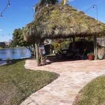 Thatched roof gazebo by a lake with green lawn and paved walkway.