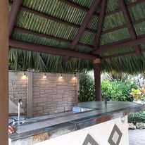 Outdoor tiki bar with thatched roof, wooden bar and stools, bottles on shelves.