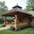 Wooden gazebo with thatched roof on a home's deck, surrounded by greenery.