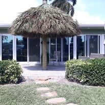 A thatch umbrella on a patio with a building in the background and green bushes and grass.