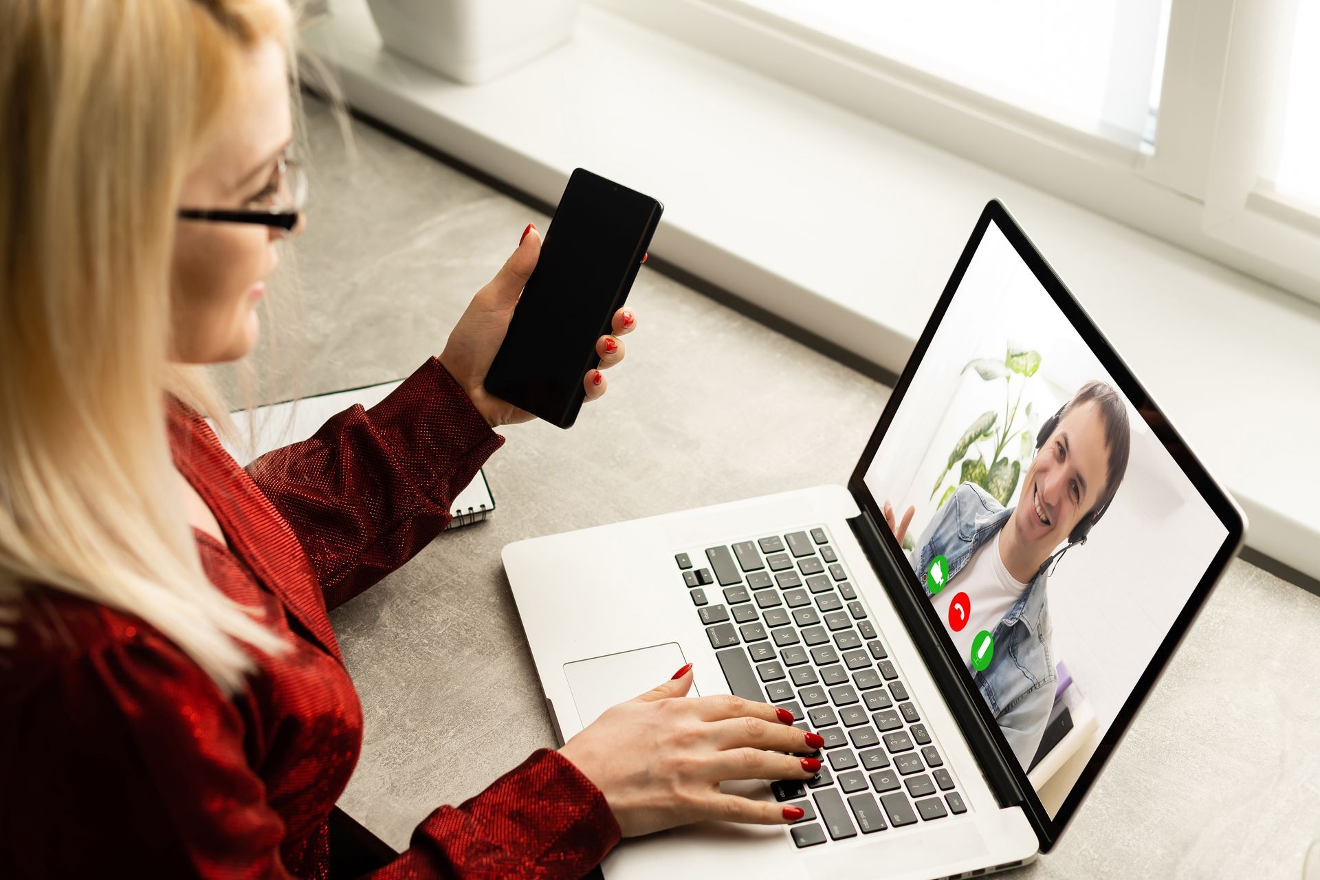 A woman is using a laptop computer while holding a cell phone.