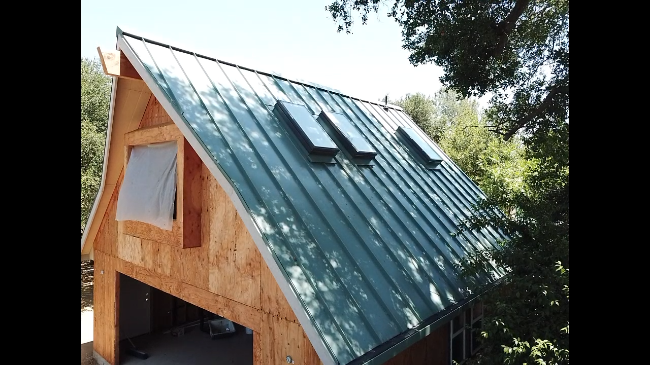 A-frame building with a green metal roof and three skylights, set against a backdrop of trees and a blue sky.