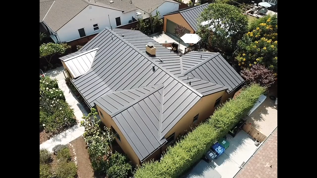 Overhead view of a house with a dark metal roof, surrounded by lush greenery and other houses in the background.