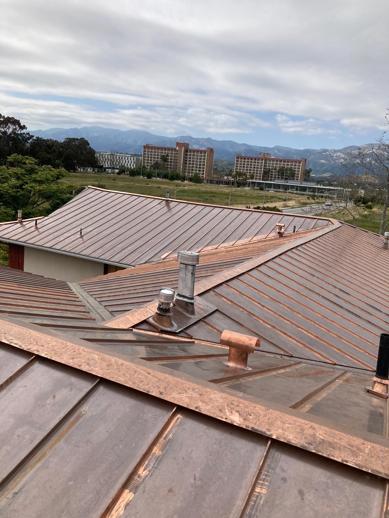 Copper roof of a building with a scenic view of other buildings and mountains under a cloudy sky.