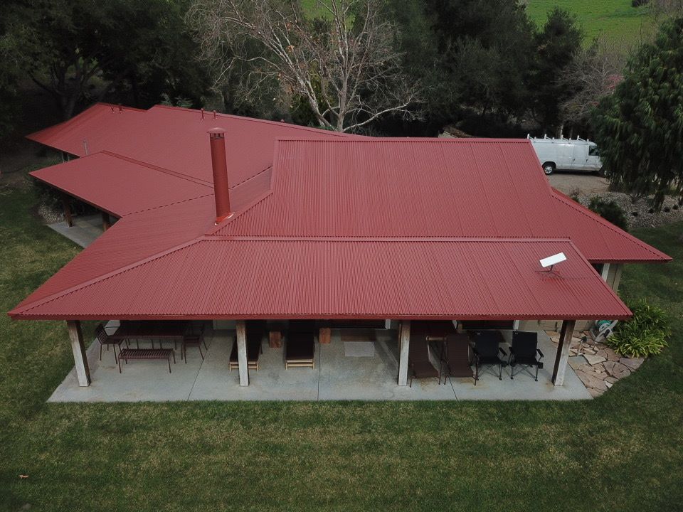 Red metal roof on a house with a porch and surrounding green grass and trees.