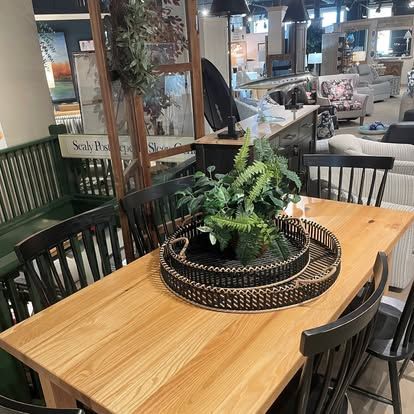 Wooden dining table with black chairs, topped with a decorative tray and plants. Interior shot of a furniture store.