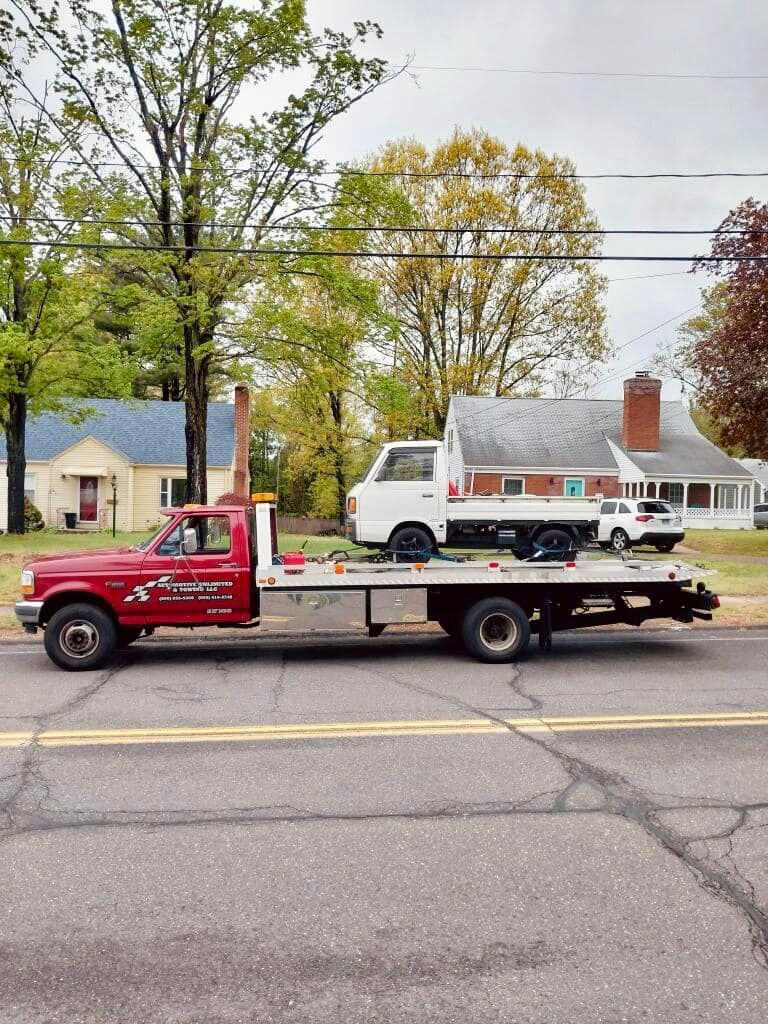 Red tow truck carrying a white vehicle on a flatbed, parked on a road in front of houses.