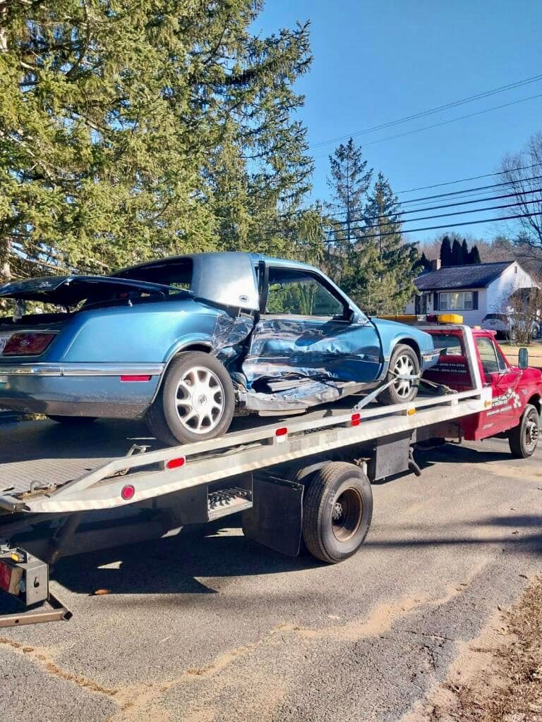 Blue car with significant damage being towed on a flatbed truck on a sunny day.