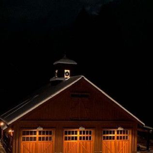 A large wooden barn with three garage doors is lit up at night.