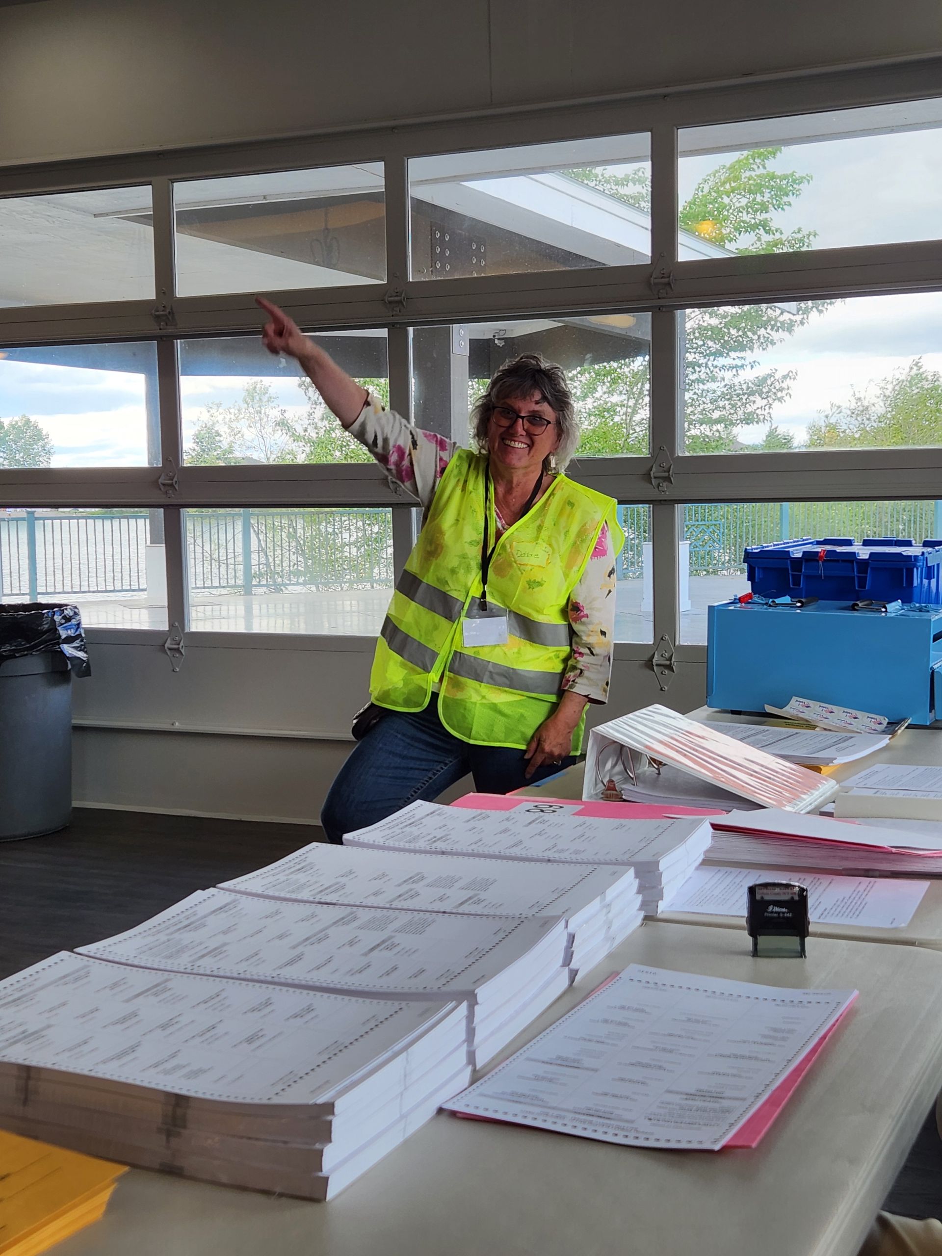 A woman in a yellow vest is sitting at a table with stacks of papers.