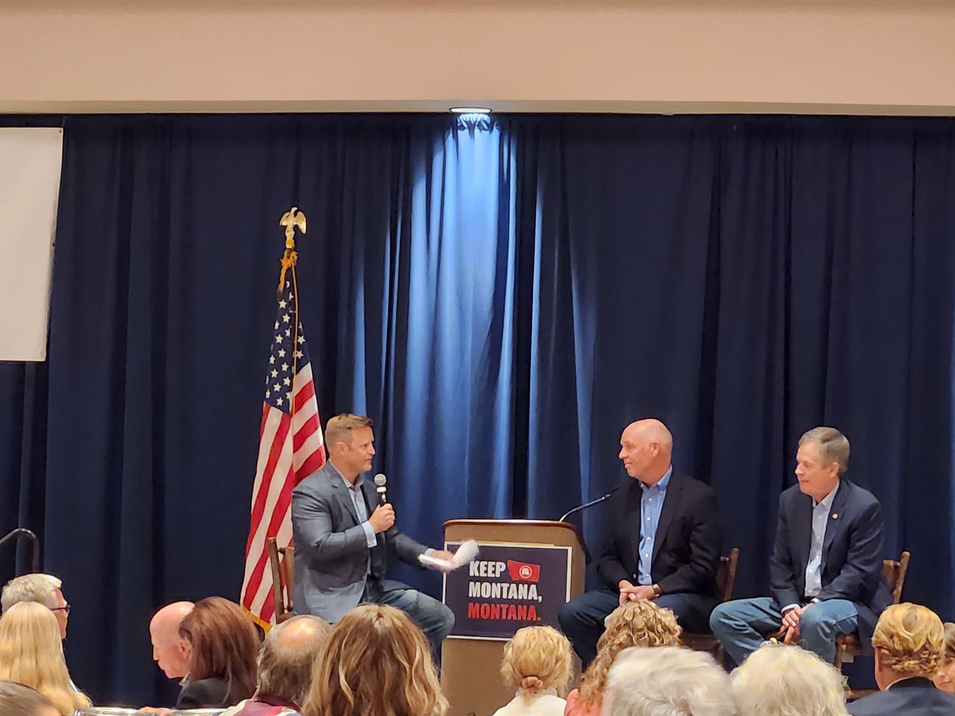Three men are sitting at a podium talking to a crowd.