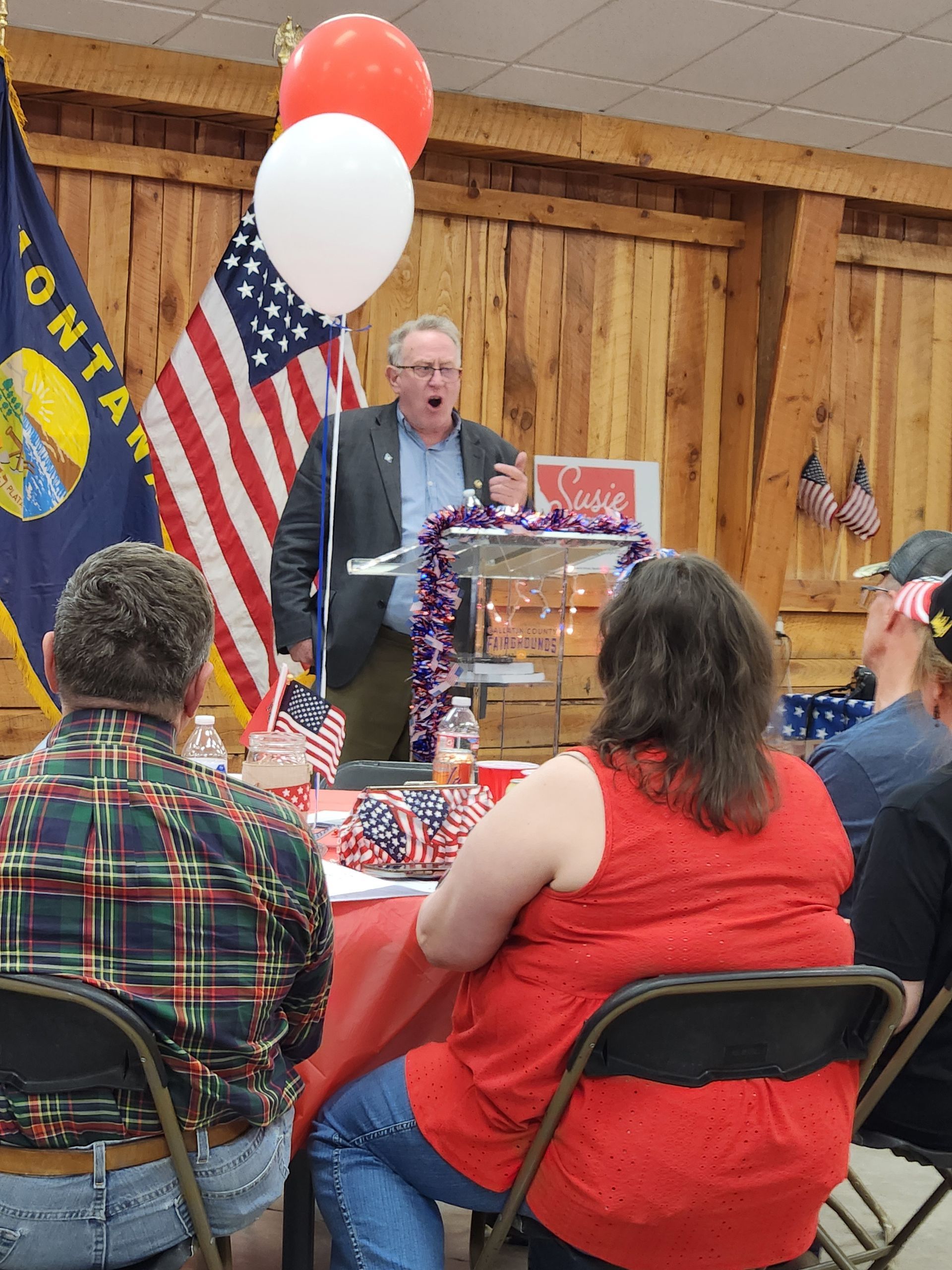 A man is giving a speech to a group of people in front of a mountain state flag