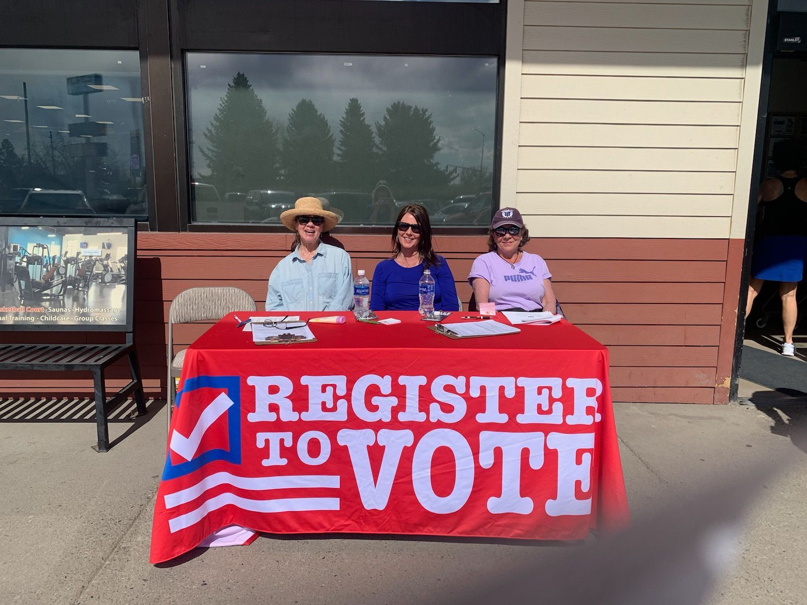 Three women sit at a table with a sign that says register to vote