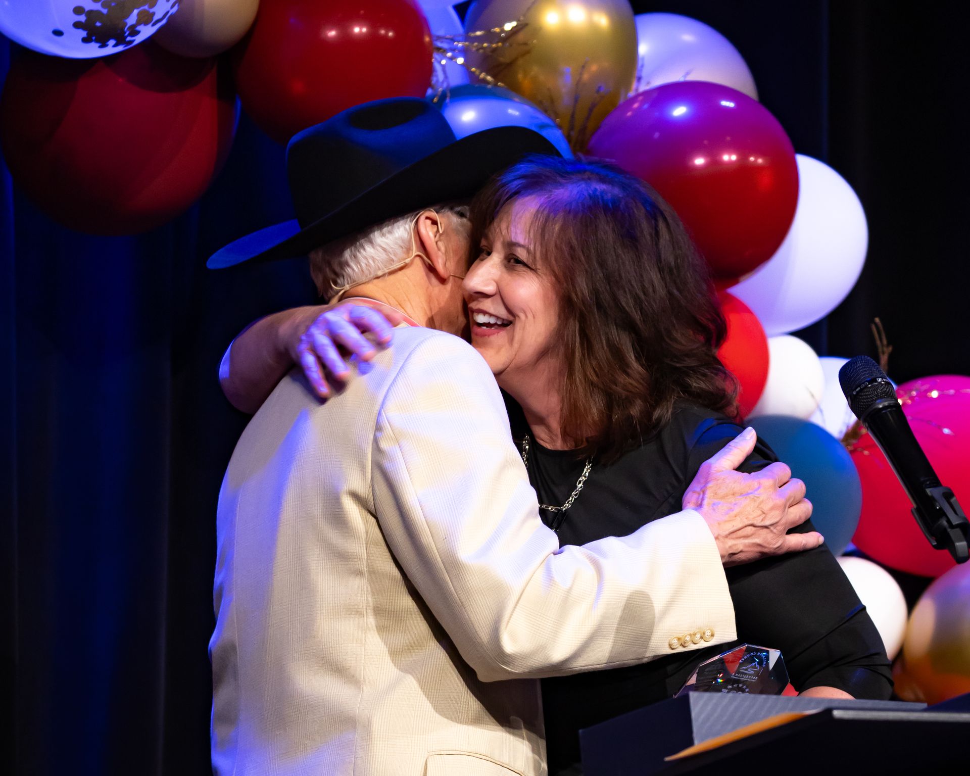 A man and woman are hugging on a stage with balloons in the background.